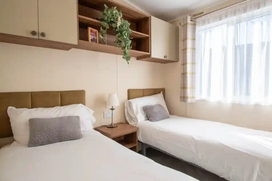 A bedroom featuring two beds with fold headboards and crisp white linen, overhead storage and a bedside table. There is a window providing natural light.