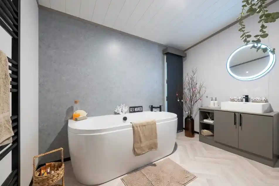 A modern bathroom featuring a freestanding white bathtub, a round illuminated mirror, and a gray accent wall. There are plants and decorative elements like a woven basket and a vase with branches. The flooring is light wood with a herringbone pattern.