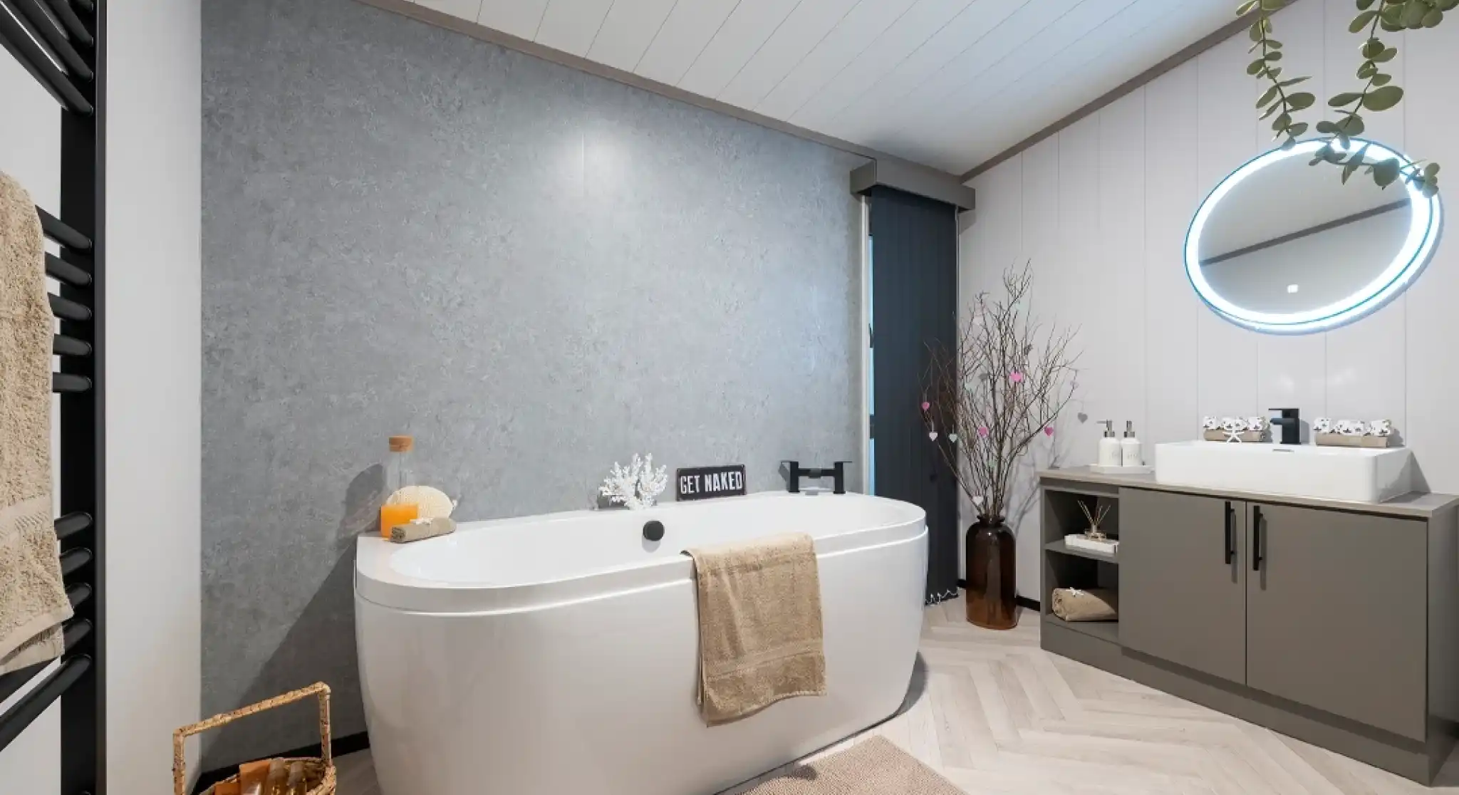 A modern bathroom featuring a freestanding white bathtub, a round illuminated mirror, and a gray accent wall. There are plants and decorative elements like a woven basket and a vase with branches. The flooring is light wood with a herringbone pattern.