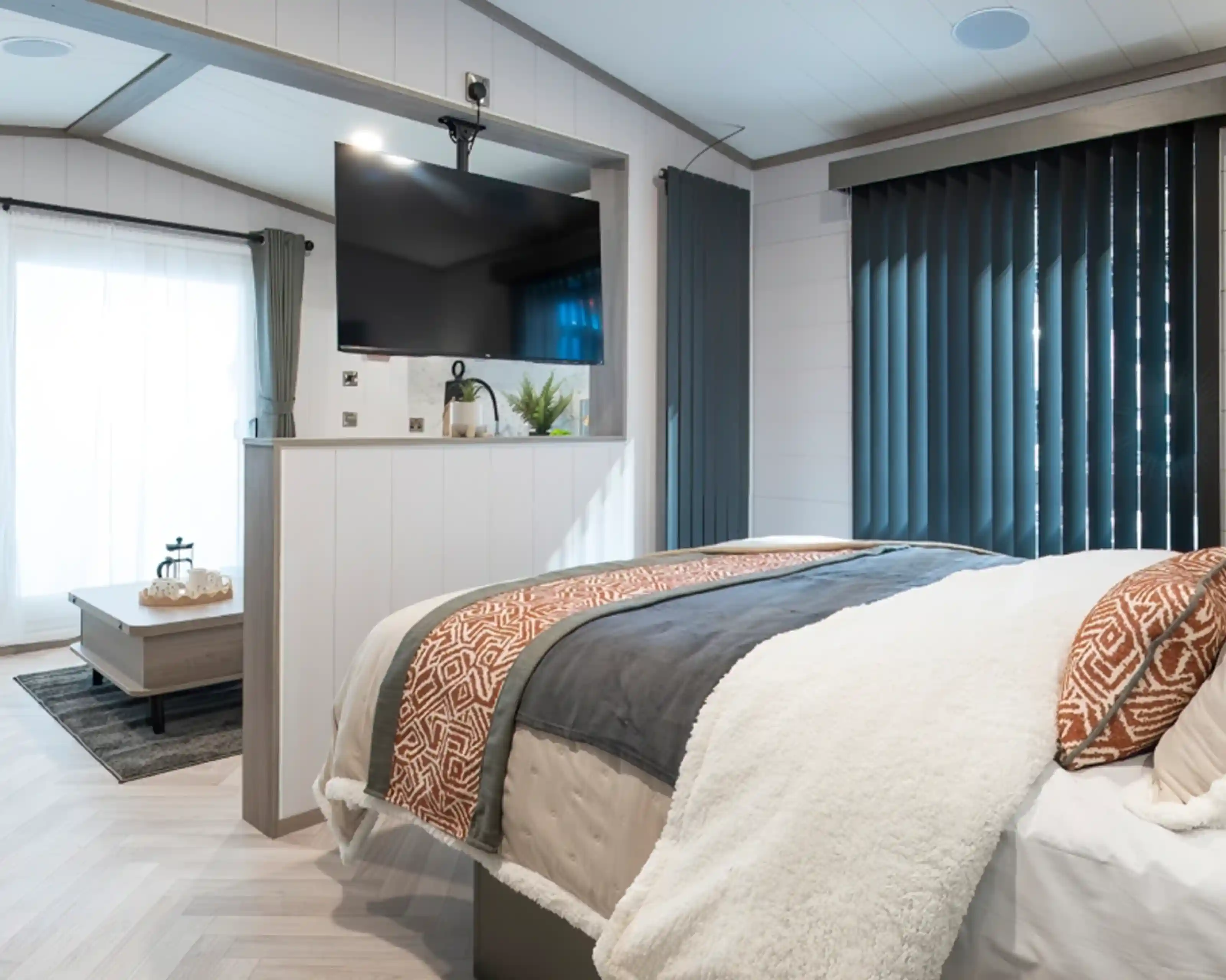 A cozy bedroom featuring a modern bed with textured throws, a large TV mounted on the wall, and tall, dark blinds. A small sitting area with a coffee table and a plant is visible in the background, along with large windows letting in natural light.