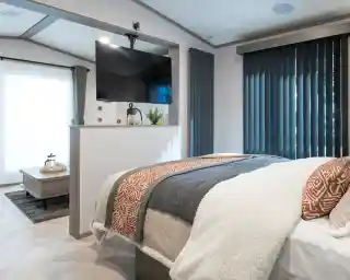 A cozy bedroom featuring a modern bed with textured throws, a large TV mounted on the wall, and tall, dark blinds. A small sitting area with a coffee table and a plant is visible in the background, along with large windows letting in natural light.