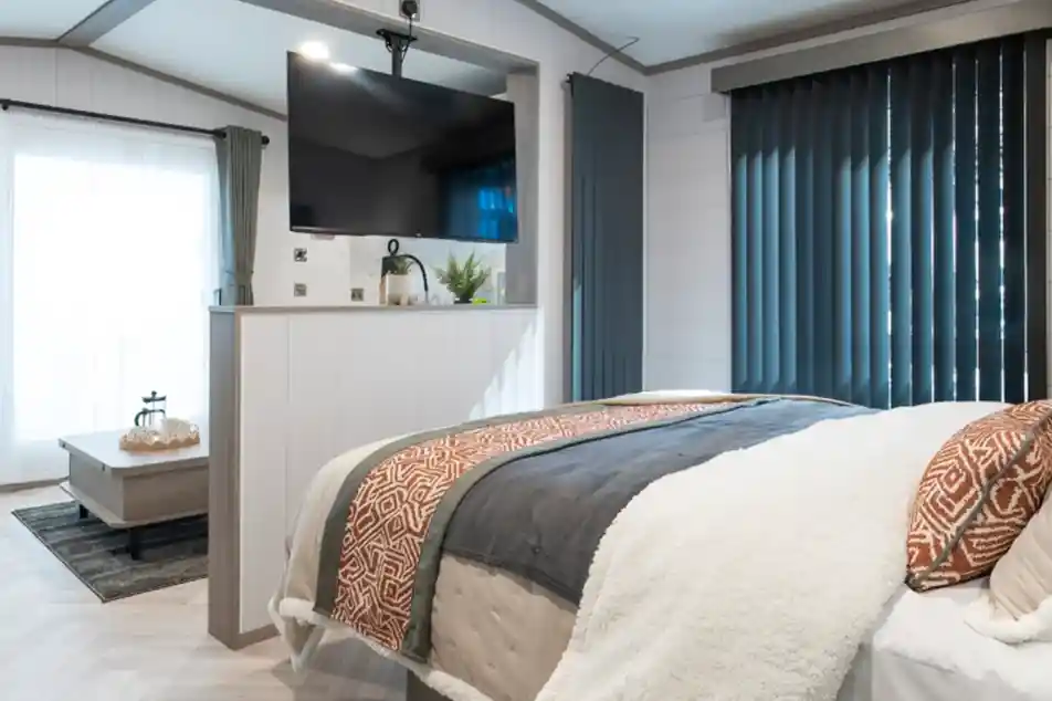 A cozy bedroom featuring a modern bed with textured throws, a large TV mounted on the wall, and tall, dark blinds. A small sitting area with a coffee table and a plant is visible in the background, along with large windows letting in natural light.