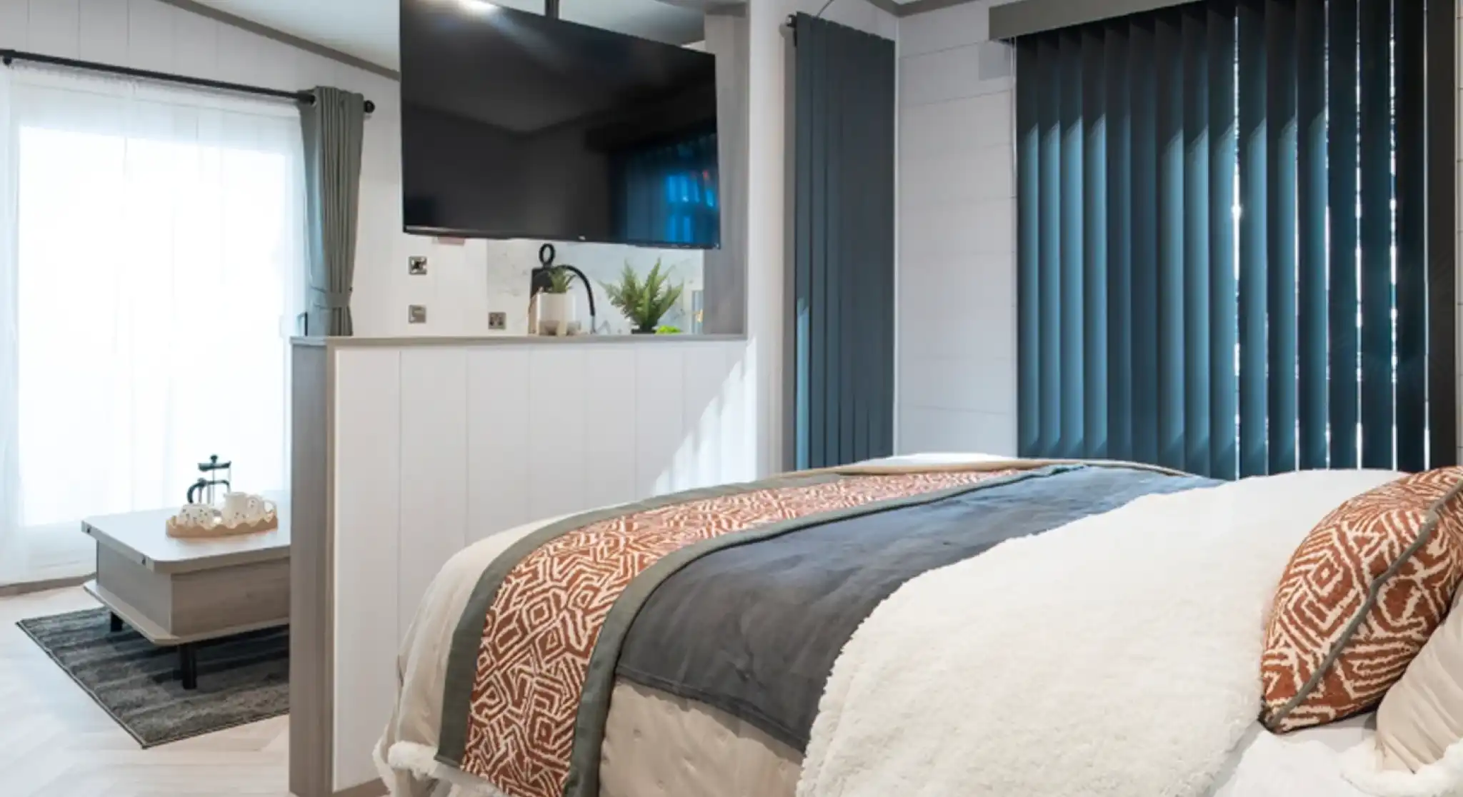 A cozy bedroom featuring a modern bed with textured throws, a large TV mounted on the wall, and tall, dark blinds. A small sitting area with a coffee table and a plant is visible in the background, along with large windows letting in natural light.