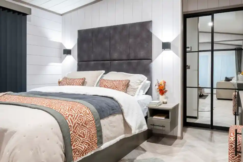 A modern bedroom featuring a plush bed with decorative pillows and a textured headboard. The walls are a light color, and the floor is designed with herringbone patterns. Soft lighting complements the cozy atmosphere, and a sliding glass door reveals a view of a living area. A small vase with orange flowers is placed on the nightstand.