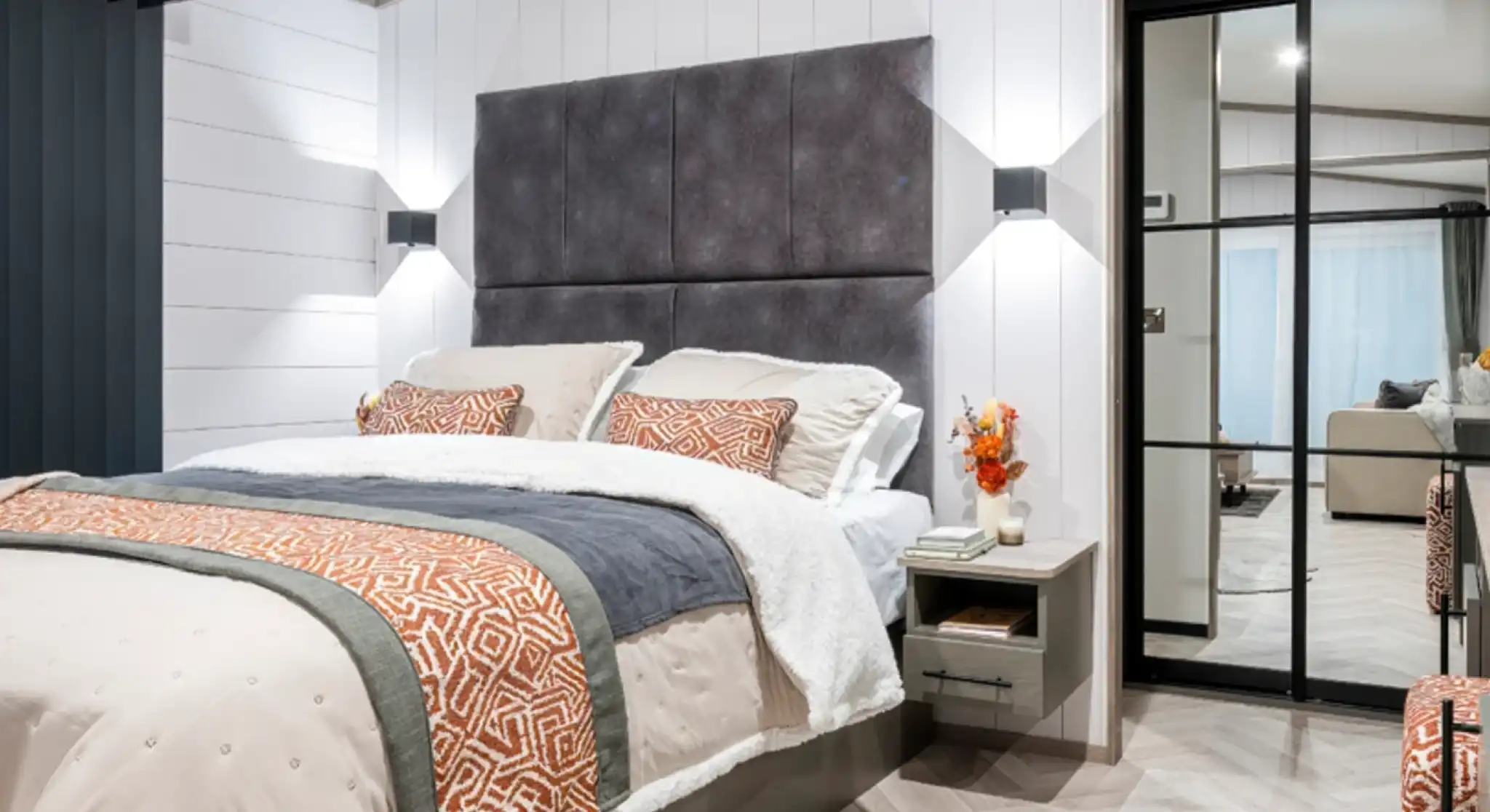 A modern bedroom featuring a plush bed with decorative pillows and a textured headboard. The walls are a light color, and the floor is designed with herringbone patterns. Soft lighting complements the cozy atmosphere, and a sliding glass door reveals a view of a living area. A small vase with orange flowers is placed on the nightstand.