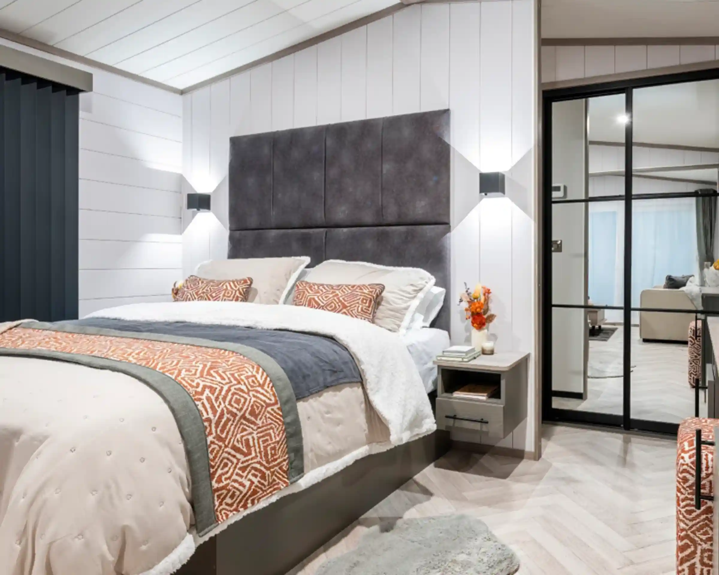 A modern bedroom featuring a plush bed with decorative pillows and a textured headboard. The walls are a light color, and the floor is designed with herringbone patterns. Soft lighting complements the cozy atmosphere, and a sliding glass door reveals a view of a living area. A small vase with orange flowers is placed on the nightstand.