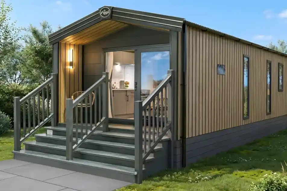 A modern, wooden-clad cabin with a slanted roof, featuring a welcoming front porch with steps. The cabin has large windows, allowing natural light to enter, and is surrounded by greenery.