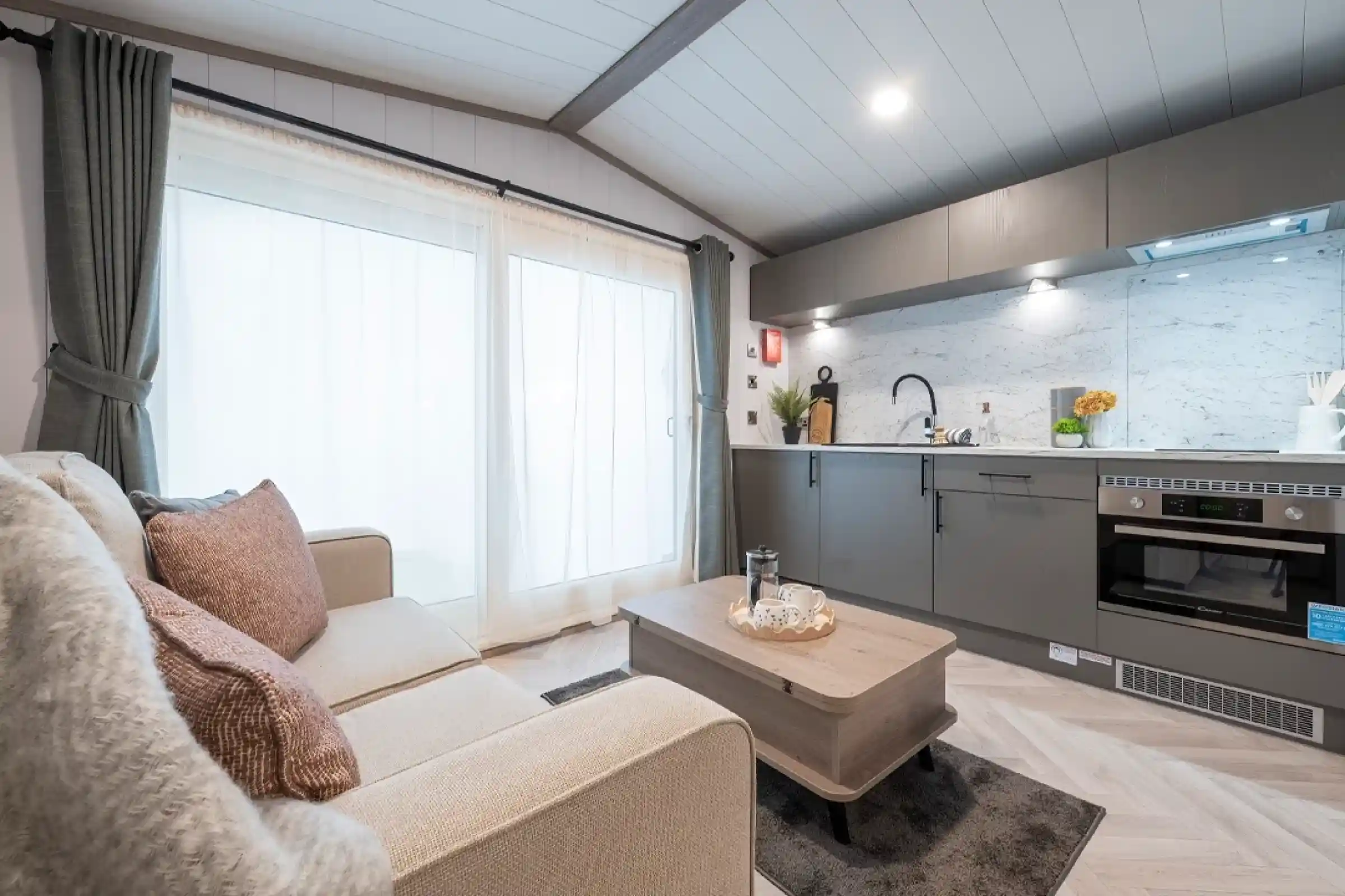 A cozy modern living space featuring a light gray sofa adorned with decorative pillows, a wooden coffee table, and a sleek kitchen with marble countertops and contemporary appliances. Large windows with sheer curtains allow natural light to fill the room.