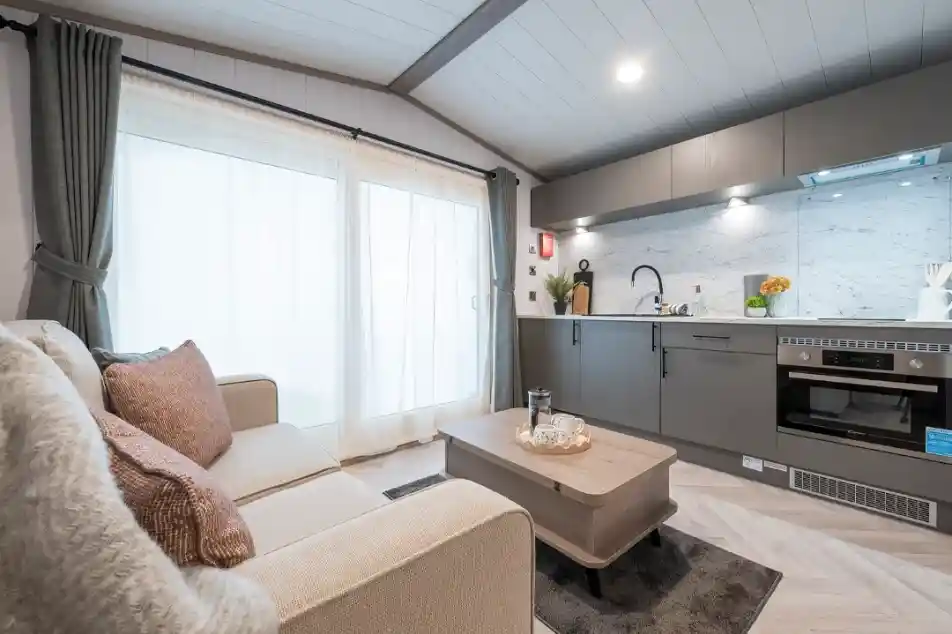 A cozy modern living space featuring a light gray sofa adorned with decorative pillows, a wooden coffee table, and a sleek kitchen with marble countertops and contemporary appliances. Large windows with sheer curtains allow natural light to fill the room.