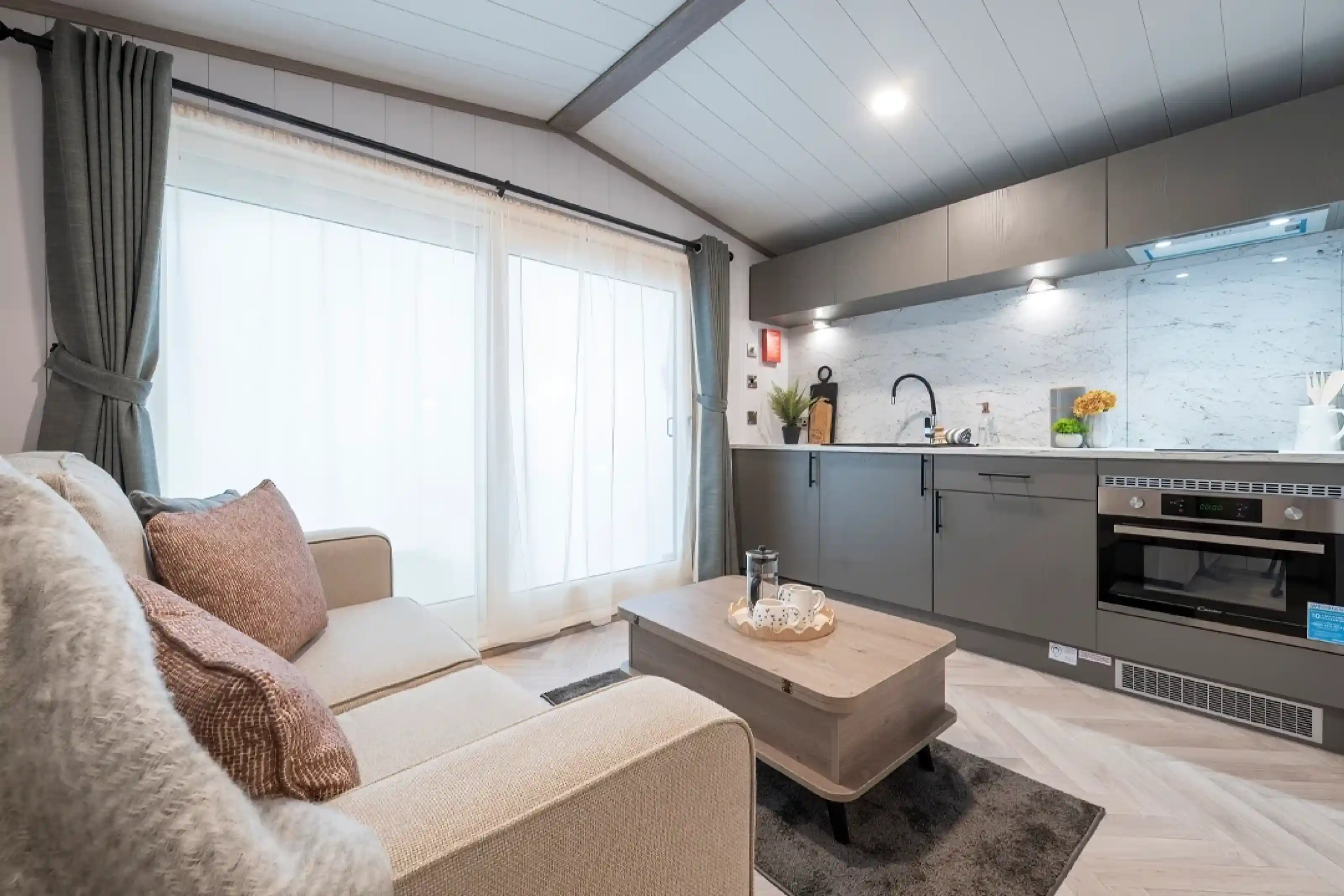 A cozy modern living space featuring a light gray sofa adorned with decorative pillows, a wooden coffee table, and a sleek kitchen with marble countertops and contemporary appliances. Large windows with sheer curtains allow natural light to fill the room.
