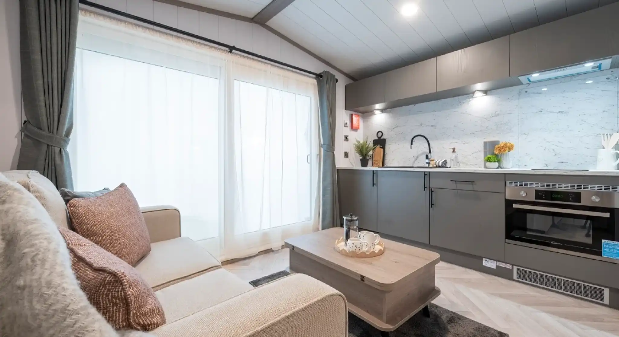 A cozy modern living space featuring a light gray sofa adorned with decorative pillows, a wooden coffee table, and a sleek kitchen with marble countertops and contemporary appliances. Large windows with sheer curtains allow natural light to fill the room.