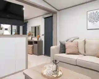 A cozy living room featuring a light-colored sofa with decorative pillows, a wooden table with a lantern and teacups, and a TV mounted on the wall. There are modern grey curtains and a dresser with decorative items in the background, creating a warm and inviting atmosphere.
