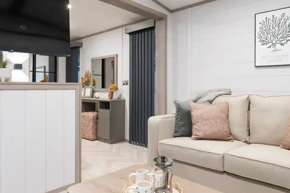 A cozy living room featuring a light-colored sofa with decorative pillows, a wooden table with a lantern and teacups, and a TV mounted on the wall. There are modern grey curtains and a dresser with decorative items in the background, creating a warm and inviting atmosphere.