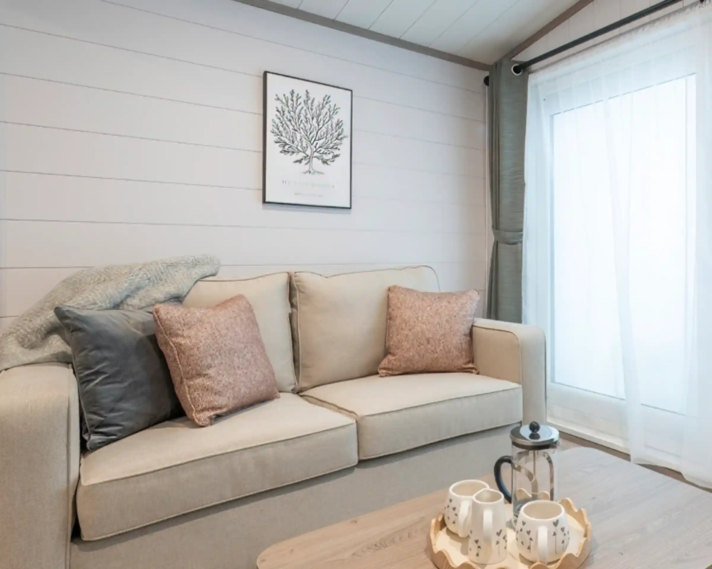 A cozy living room featuring a light-colored sofa with decorative pillows, a wooden coffee table with tea set, and a large window with soft curtains. A botanical print hangs on the wall, complementing the neutral color scheme.