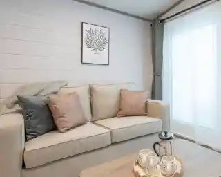 A cozy living room featuring a light-colored sofa with decorative pillows, a wooden coffee table with tea set, and a large window with soft curtains. A botanical print hangs on the wall, complementing the neutral color scheme.