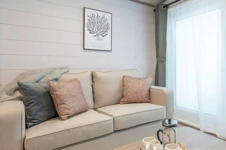 A cozy living room featuring a light-colored sofa with decorative pillows, a wooden coffee table with tea set, and a large window with soft curtains. A botanical print hangs on the wall, complementing the neutral color scheme.