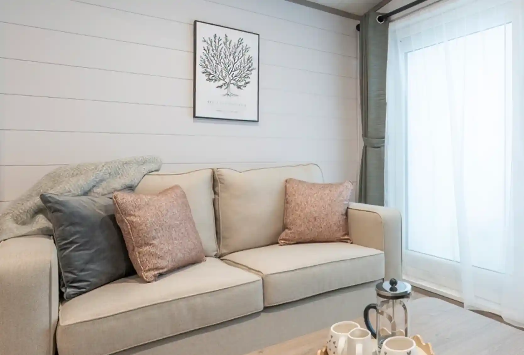 A cozy living room featuring a light-colored sofa with decorative pillows, a wooden coffee table with tea set, and a large window with soft curtains. A botanical print hangs on the wall, complementing the neutral color scheme.