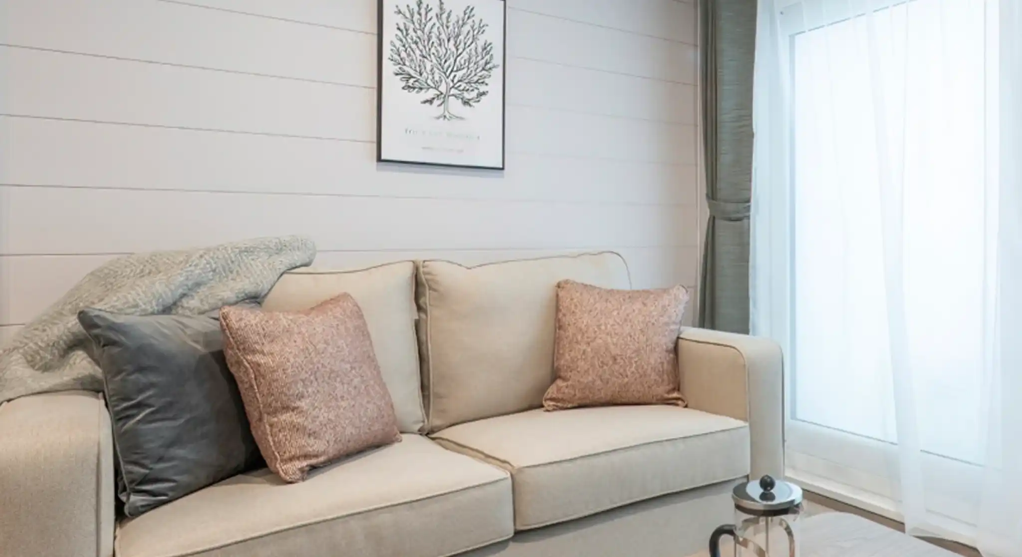 A cozy living room featuring a light-colored sofa with decorative pillows, a wooden coffee table with tea set, and a large window with soft curtains. A botanical print hangs on the wall, complementing the neutral color scheme.