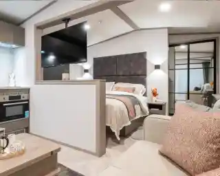 A cozy modern studio bedroom features a bed with a gray headboard, a flat-screen TV mounted on the wall, and a small kitchenette. Decorative pillows and a vase of flowers add a welcoming touch. A glass door leads to a closet, while neutral tones create a warm atmosphere.
