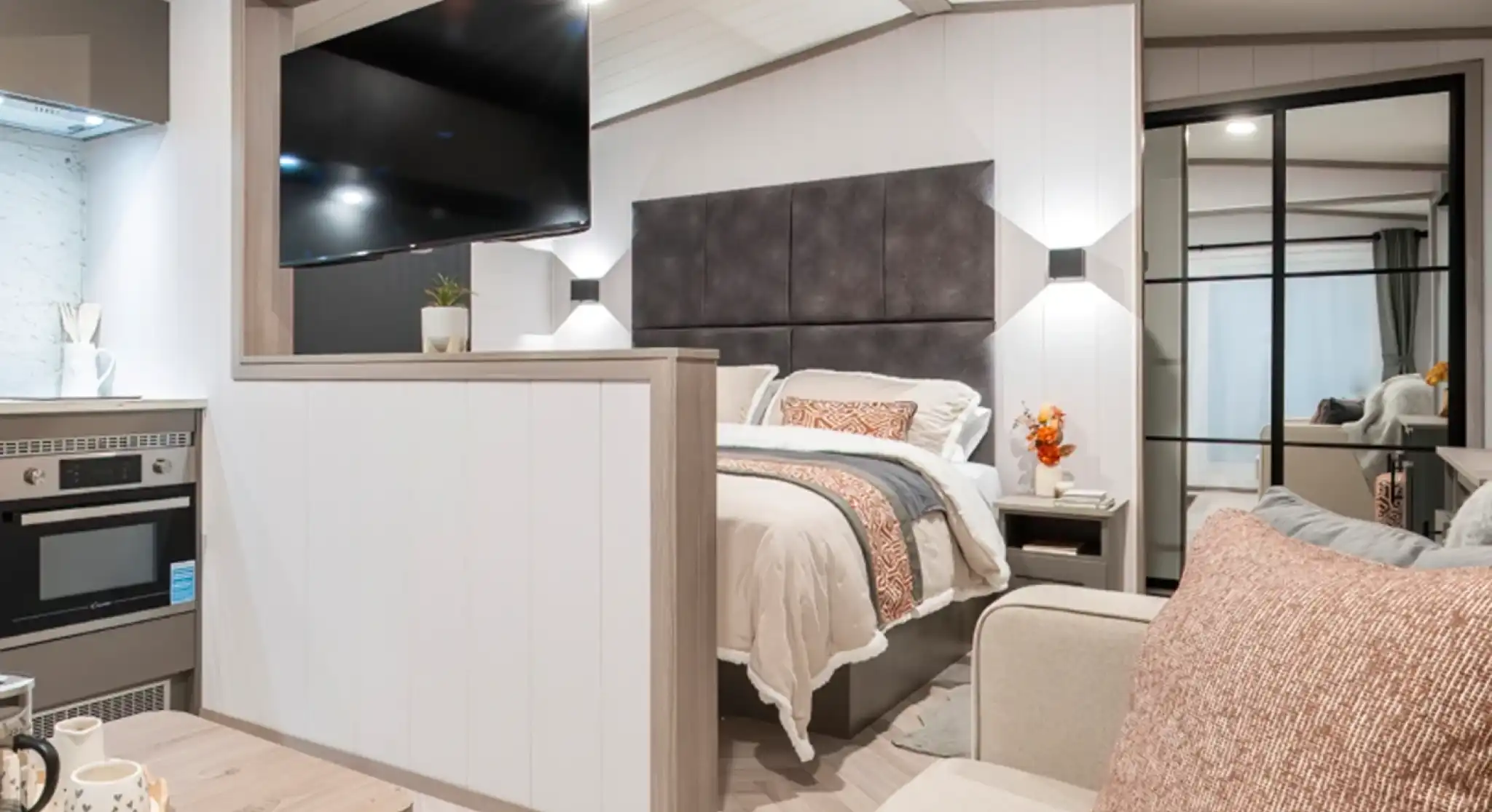 A cozy modern studio bedroom features a bed with a gray headboard, a flat-screen TV mounted on the wall, and a small kitchenette. Decorative pillows and a vase of flowers add a welcoming touch. A glass door leads to a closet, while neutral tones create a warm atmosphere.