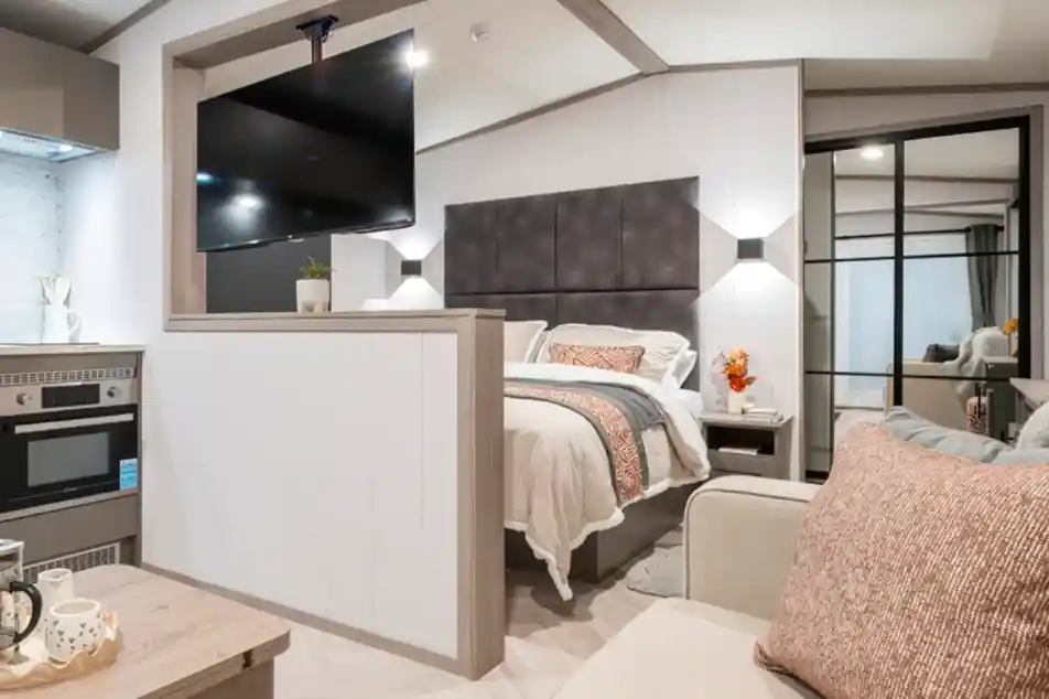 A cozy modern studio bedroom features a bed with a gray headboard, a flat-screen TV mounted on the wall, and a small kitchenette. Decorative pillows and a vase of flowers add a welcoming touch. A glass door leads to a closet, while neutral tones create a warm atmosphere.