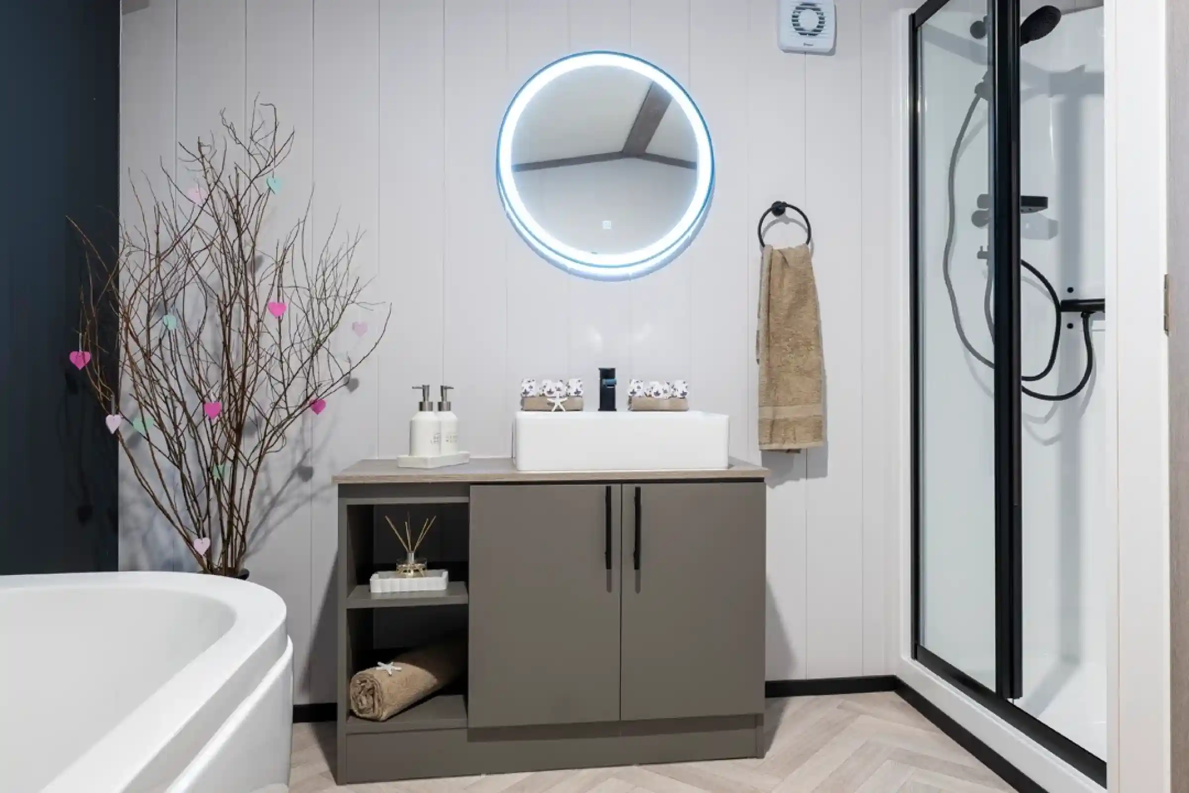 A modern bathroom featuring a round illuminated mirror above a sleek gray vanity. The vanity holds a white sink, decorative items, and neatly arranged towels. A simple tree branch with colorful hearts adds a touch of decor. A shower cubicle is visible in the corner, alongside a white freestanding bathtub.