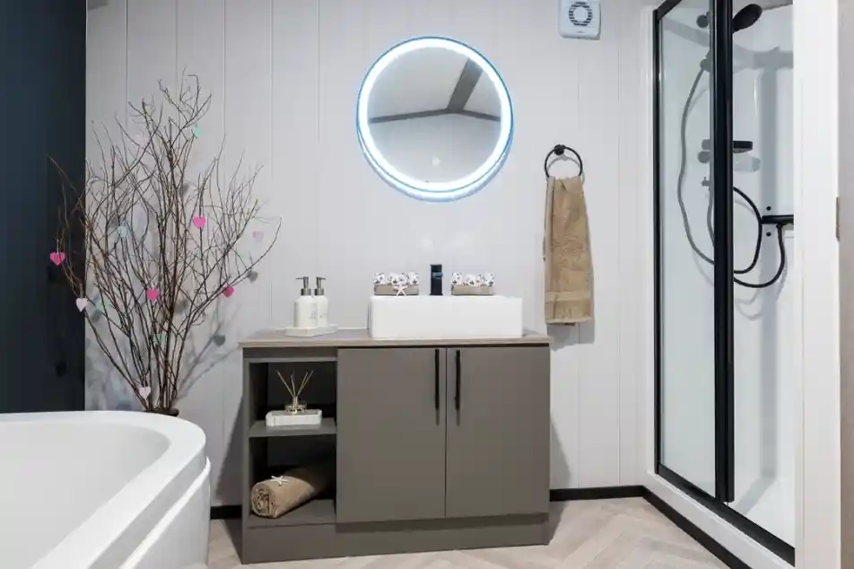 A modern bathroom featuring a round illuminated mirror above a sleek gray vanity. The vanity holds a white sink, decorative items, and neatly arranged towels. A simple tree branch with colorful hearts adds a touch of decor. A shower cubicle is visible in the corner, alongside a white freestanding bathtub.