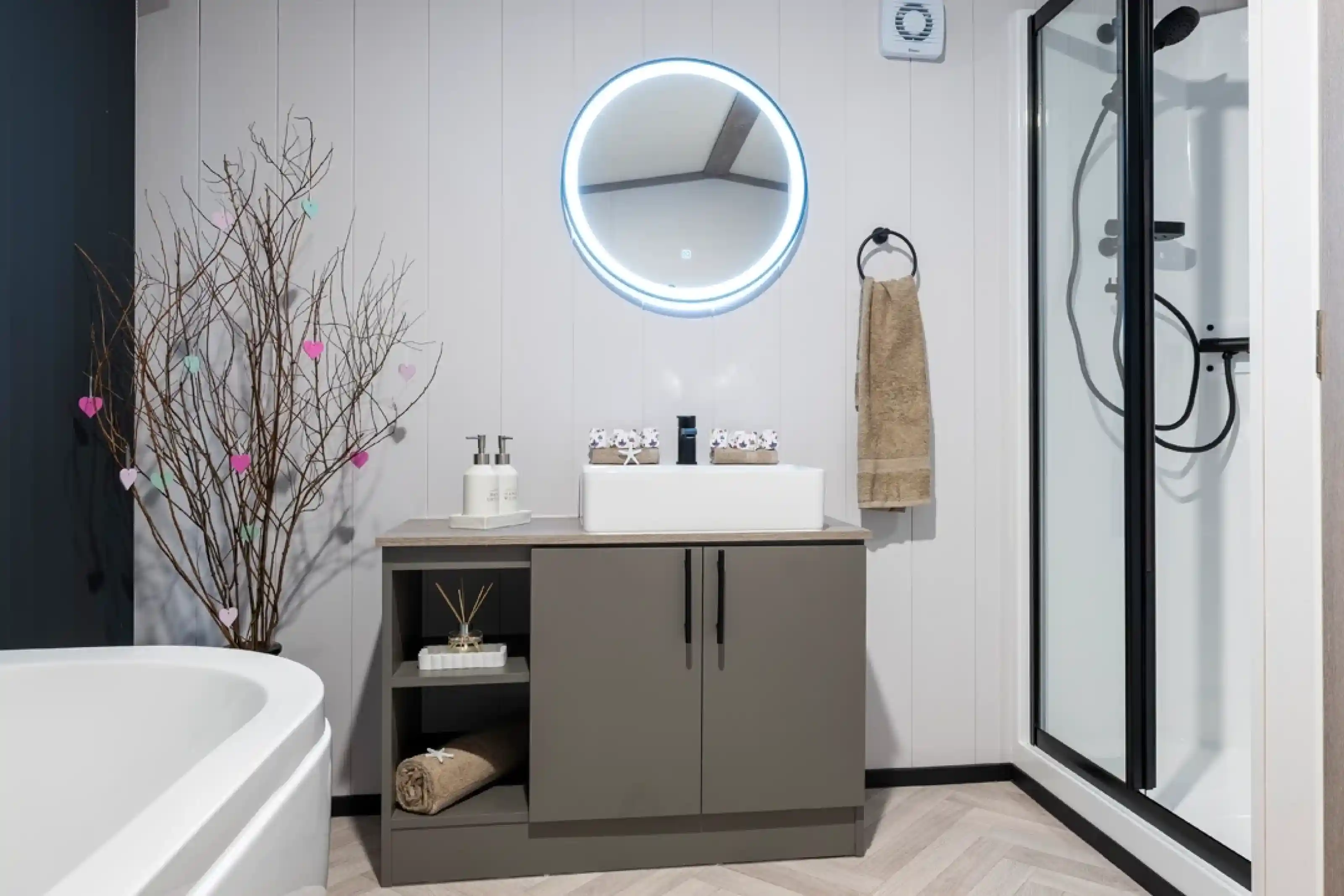 A modern bathroom featuring a round illuminated mirror above a sleek gray vanity. The vanity holds a white sink, decorative items, and neatly arranged towels. A simple tree branch with colorful hearts adds a touch of decor. A shower cubicle is visible in the corner, alongside a white freestanding bathtub.
