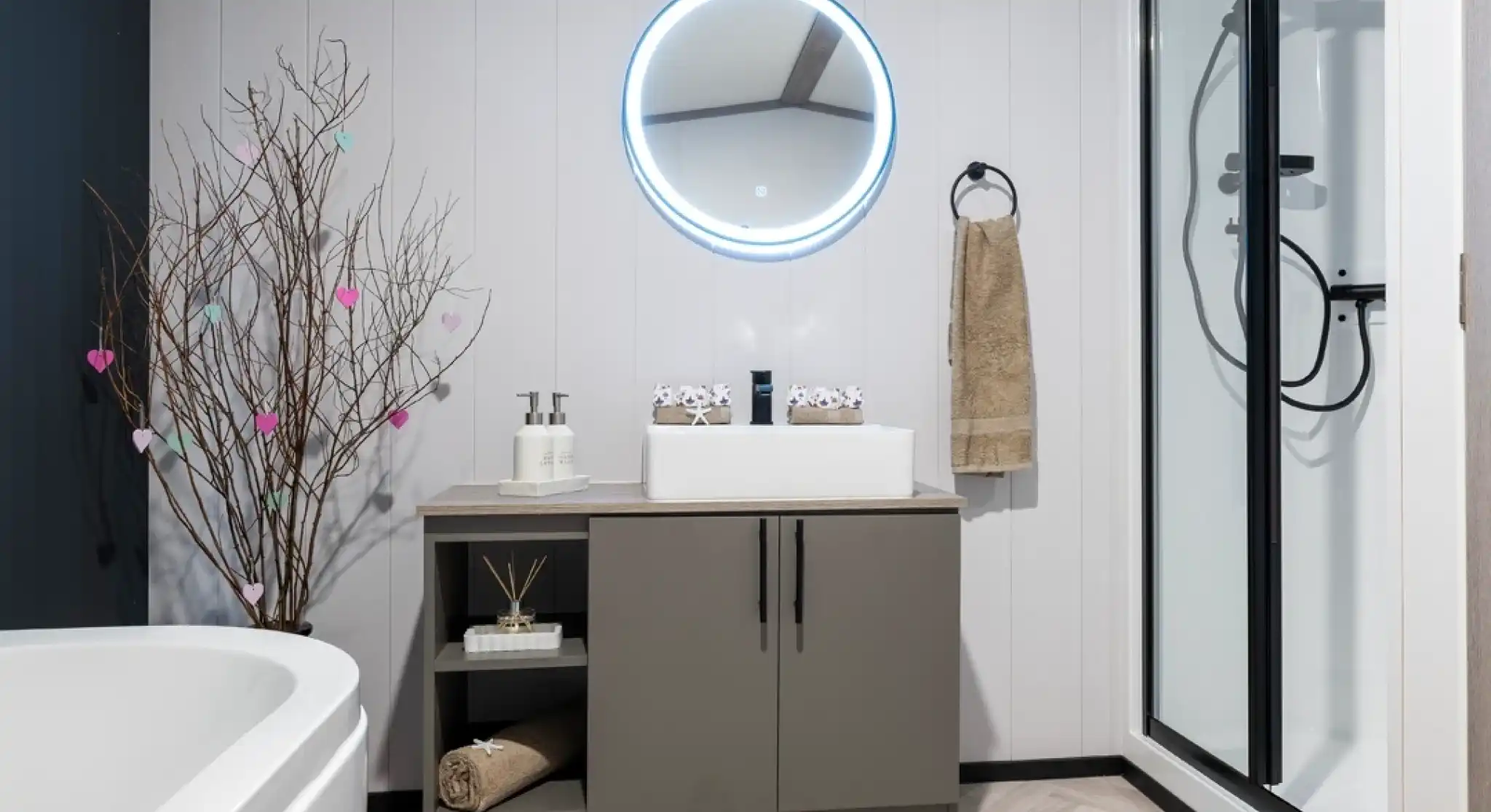 A modern bathroom featuring a round illuminated mirror above a sleek gray vanity. The vanity holds a white sink, decorative items, and neatly arranged towels. A simple tree branch with colorful hearts adds a touch of decor. A shower cubicle is visible in the corner, alongside a white freestanding bathtub.