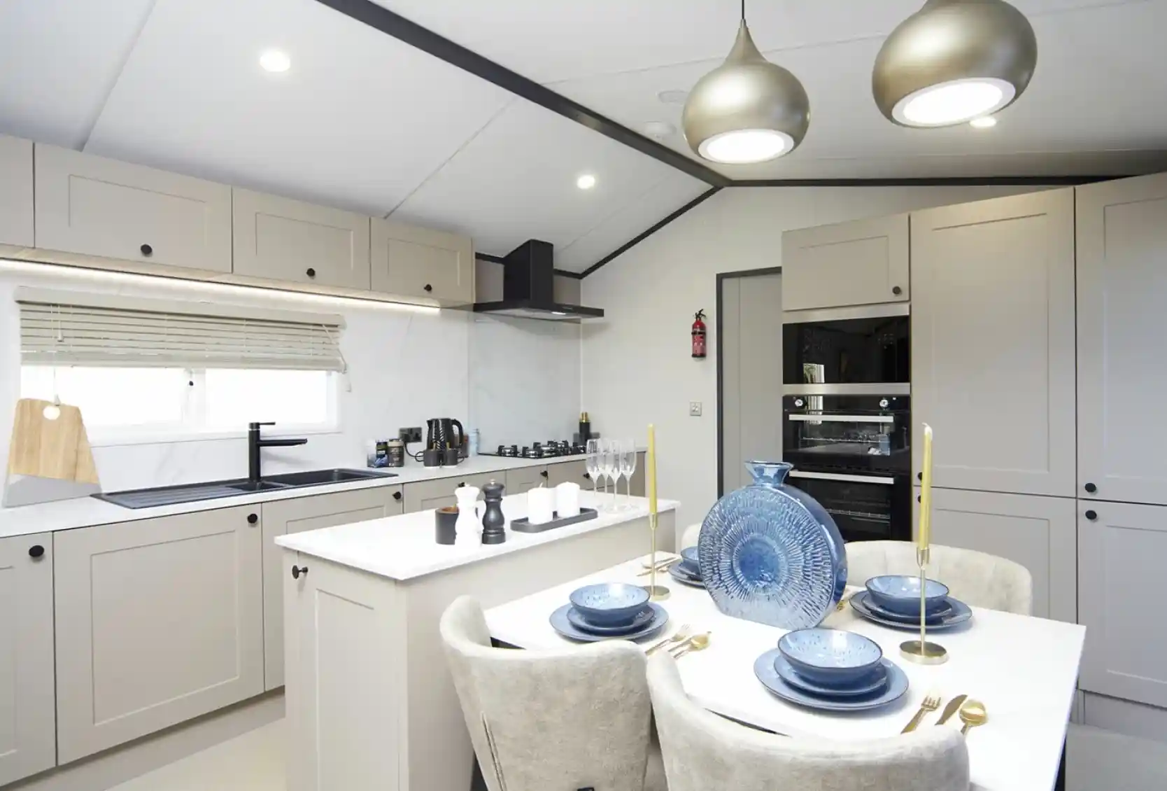 A modern kitchen-diner with light grey cabinets, a white island, and built-in oven. The dining table is set with blue plates, gold cutlery, and a decorative blue vase. Two pendant lights hang overhead, and the kitchen is bright with natural and ceiling lighting.