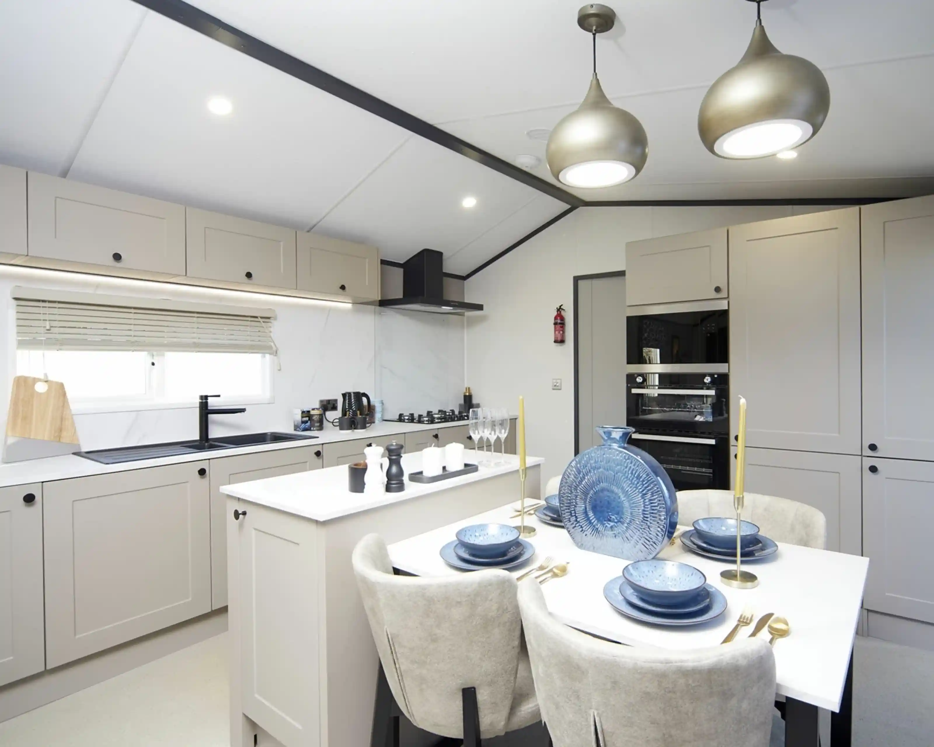 A modern kitchen-diner with light grey cabinets, a white island, and built-in oven. The dining table is set with blue plates, gold cutlery, and a decorative blue vase. Two pendant lights hang overhead, and the kitchen is bright with natural and ceiling lighting.