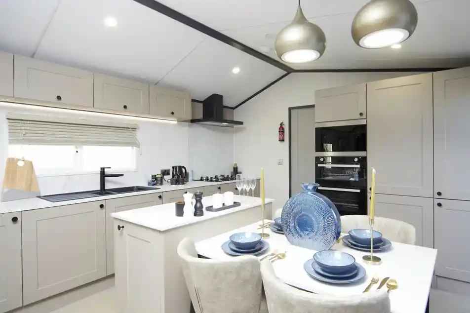 A modern kitchen-diner with light grey cabinets, a white island, and built-in oven. The dining table is set with blue plates, gold cutlery, and a decorative blue vase. Two pendant lights hang overhead, and the kitchen is bright with natural and ceiling lighting.