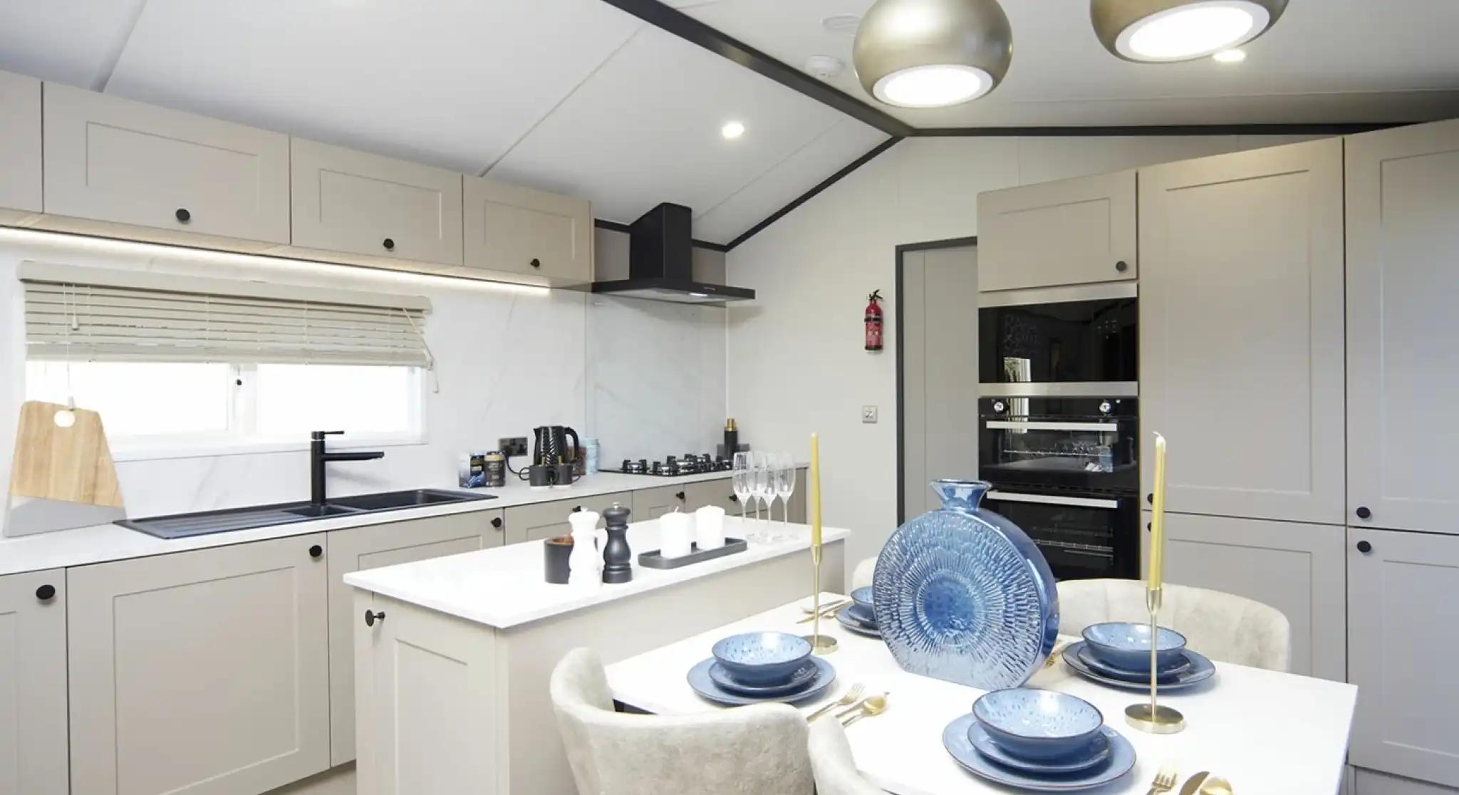 A modern kitchen-diner with light grey cabinets, a white island, and built-in oven. The dining table is set with blue plates, gold cutlery, and a decorative blue vase. Two pendant lights hang overhead, and the kitchen is bright with natural and ceiling lighting.