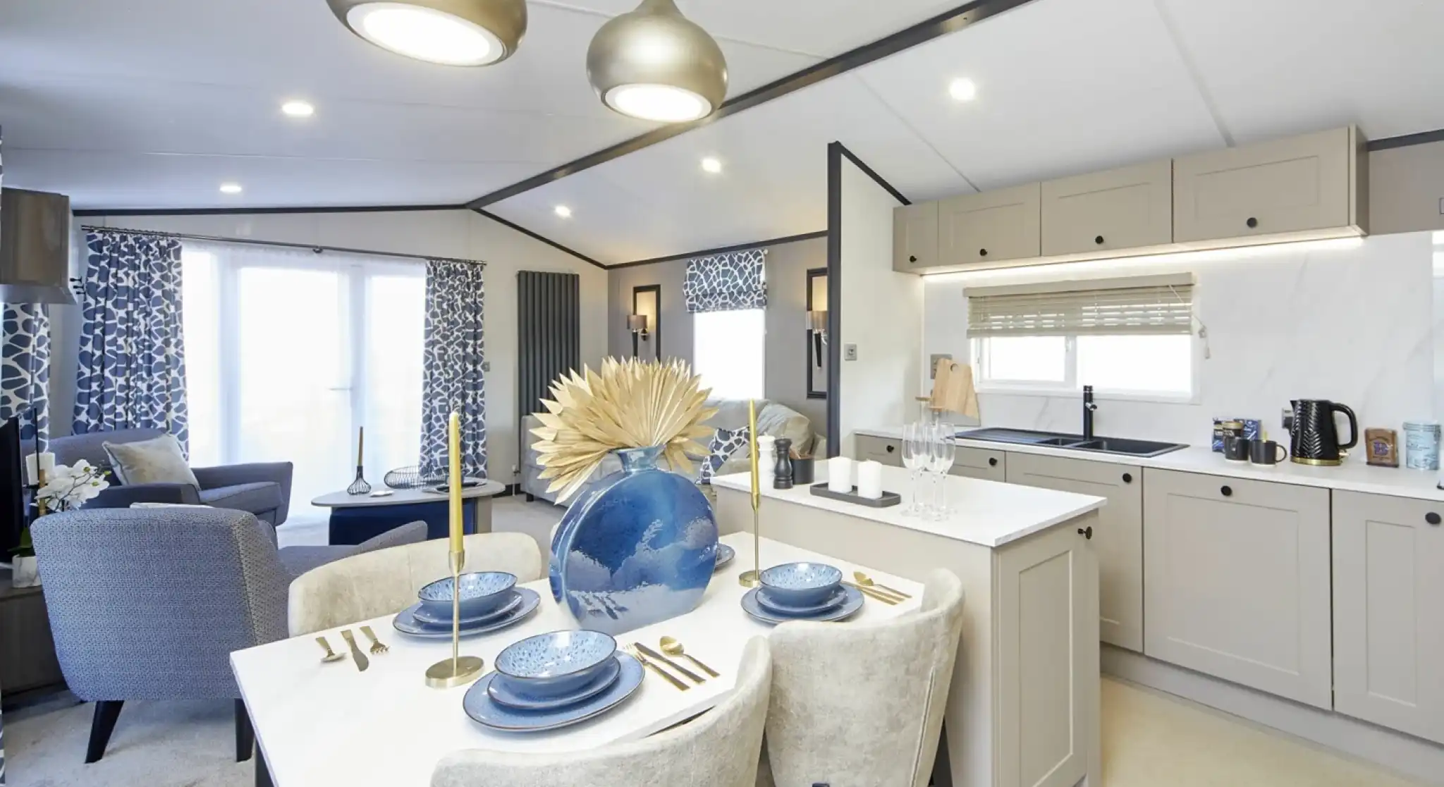 An open-plan living space with a modern kitchen and dining area leading into a lounge. The dining table is set with blue dishes and a large decorative vase, while the lounge has patterned blue curtains, blue chairs, and a bright, airy feel with large windows.