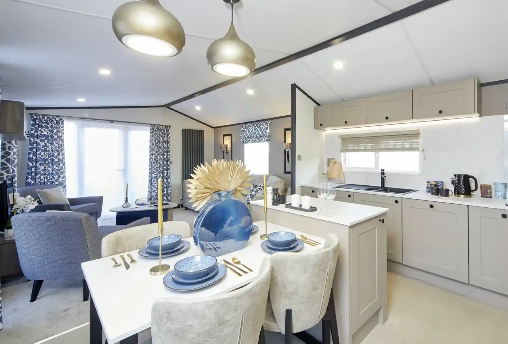 An open-plan living space with a modern kitchen and dining area leading into a lounge. The dining table is set with blue dishes and a large decorative vase, while the lounge has patterned blue curtains, blue chairs, and a bright, airy feel with large windows.