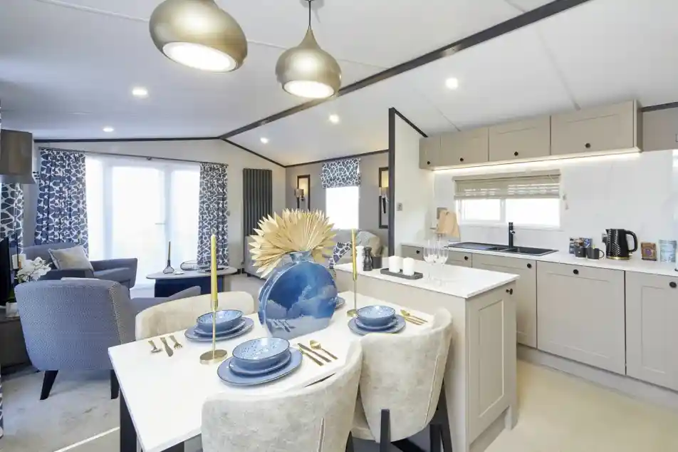 An open-plan living space with a modern kitchen and dining area leading into a lounge. The dining table is set with blue dishes and a large decorative vase, while the lounge has patterned blue curtains, blue chairs, and a bright, airy feel with large windows.