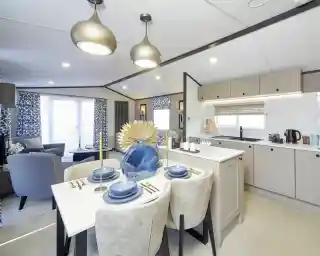 An open-plan living space with a modern kitchen and dining area leading into a lounge. The dining table is set with blue dishes and a large decorative vase, while the lounge has patterned blue curtains, blue chairs, and a bright, airy feel with large windows.