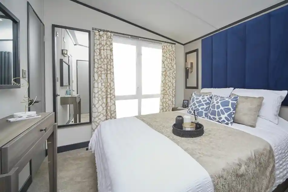 A stylish bedroom with a large bed dressed in white and beige bedding, complemented by blue patterned cushions and a tall blue headboard. A mirror and dressing table sit to the side, with patterned curtains covering floor-to-ceiling windows, and decorative items neatly arranged on the bedside table.