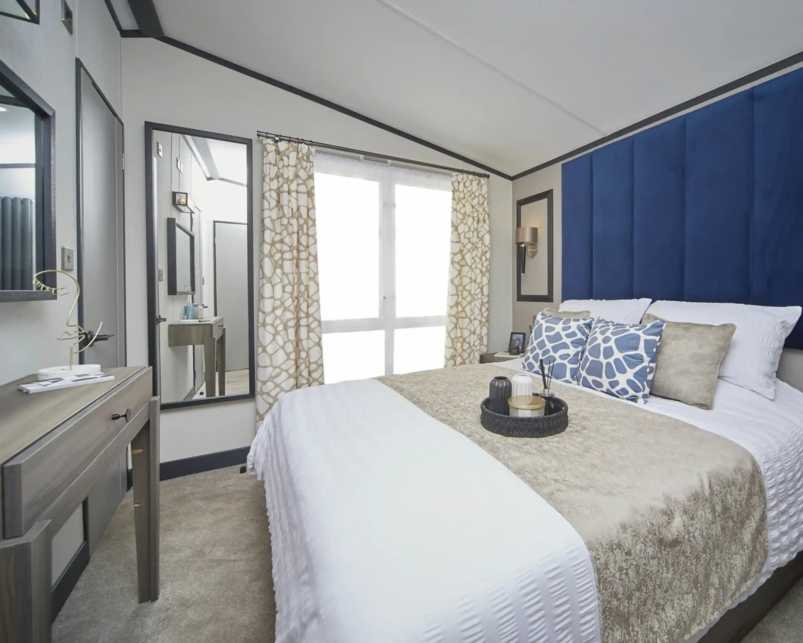 A stylish bedroom with a large bed dressed in white and beige bedding, complemented by blue patterned cushions and a tall blue headboard. A mirror and dressing table sit to the side, with patterned curtains covering floor-to-ceiling windows, and decorative items neatly arranged on the bedside table.