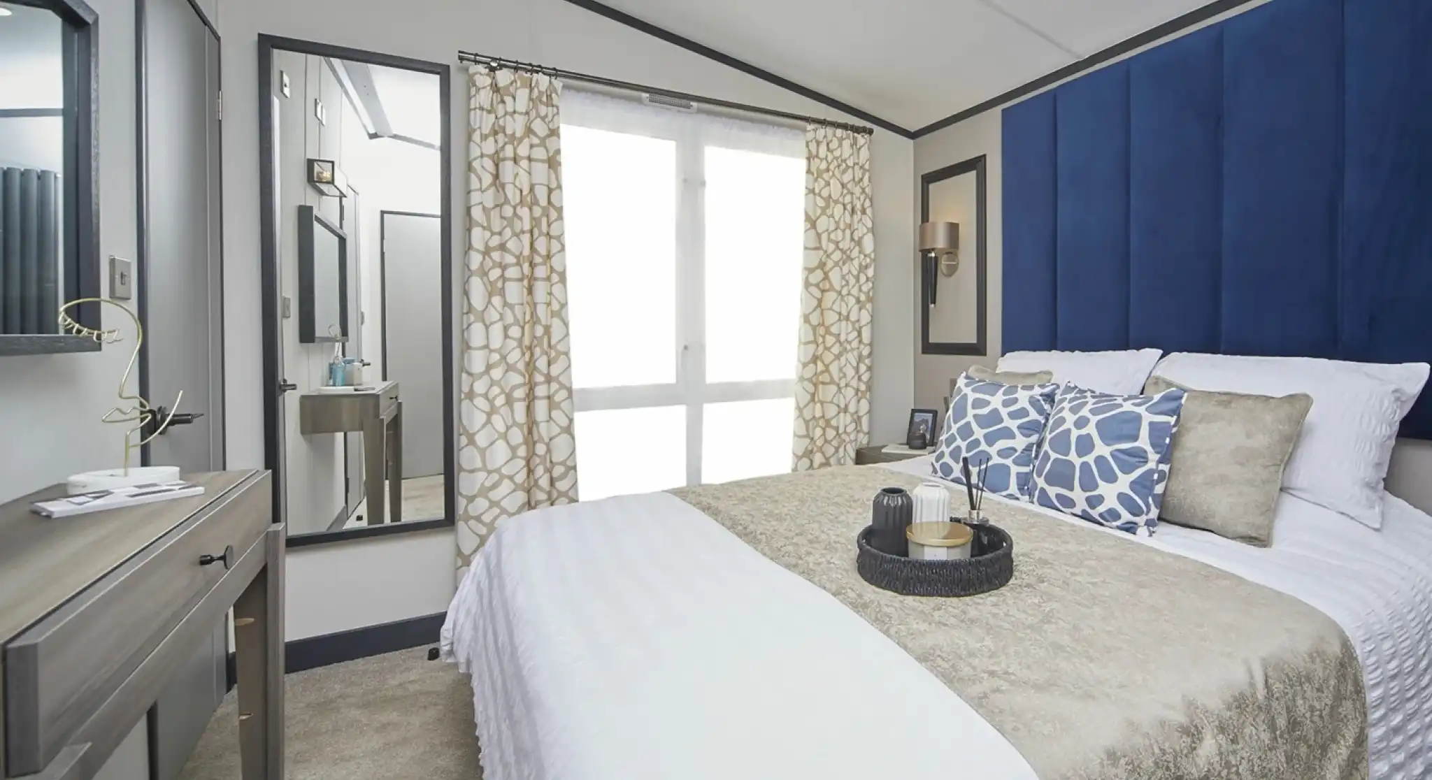 A stylish bedroom with a large bed dressed in white and beige bedding, complemented by blue patterned cushions and a tall blue headboard. A mirror and dressing table sit to the side, with patterned curtains covering floor-to-ceiling windows, and decorative items neatly arranged on the bedside table.