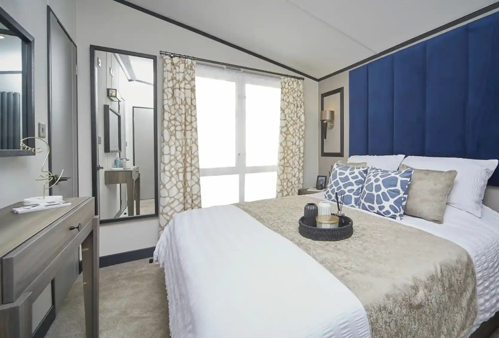 A stylish bedroom with a large bed dressed in white and beige bedding, complemented by blue patterned cushions and a tall blue headboard. A mirror and dressing table sit to the side, with patterned curtains covering floor-to-ceiling windows, and decorative items neatly arranged on the bedside table.