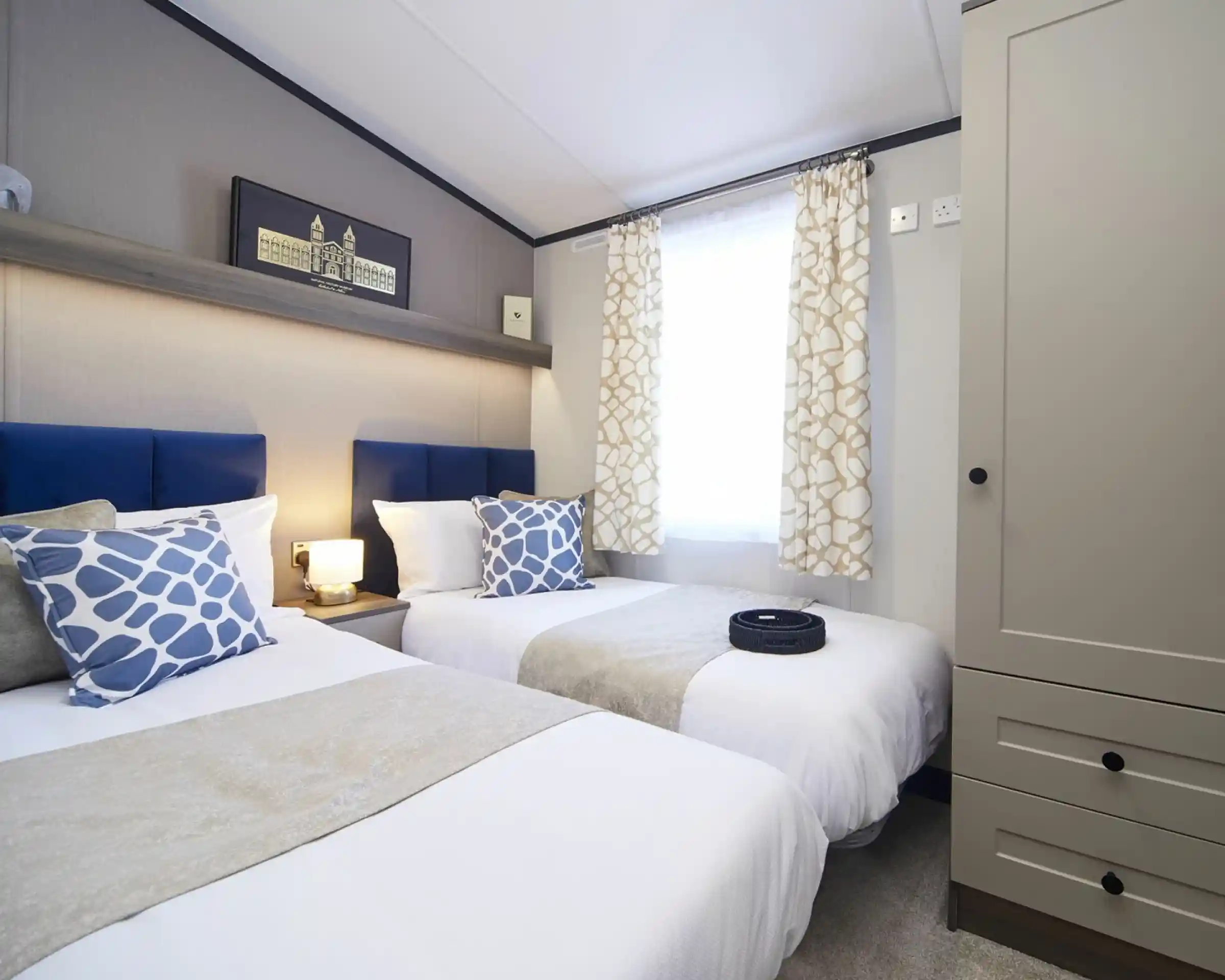 A stylish twin bedroom with twin beds with matching blue headboards, blue patterned cushions and white crisp linen. There is also a wardrobe and a window with patterned curtains allowing natural light.