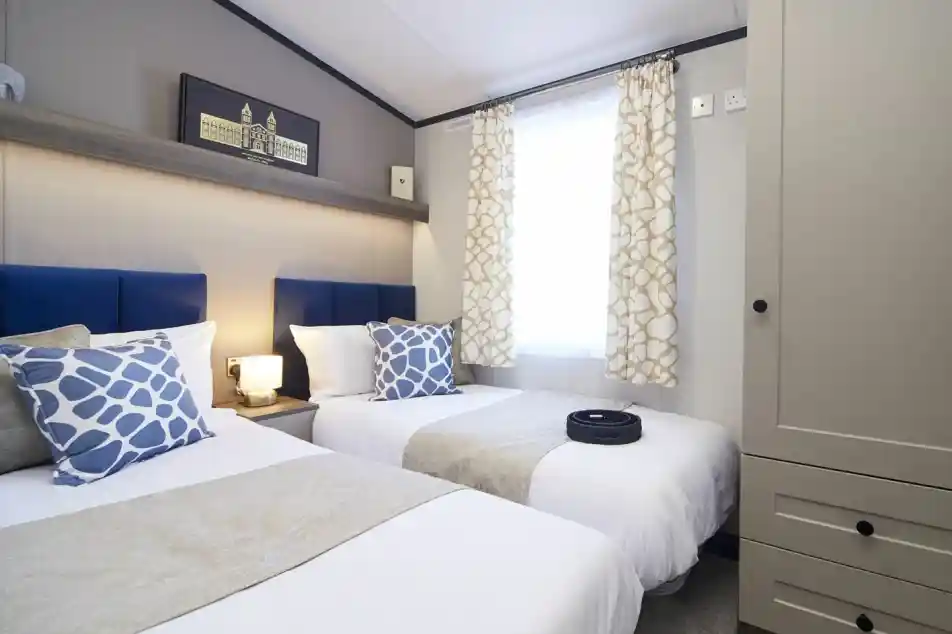 A stylish twin bedroom with twin beds with matching blue headboards, blue patterned cushions and white crisp linen. There is also a wardrobe and a window with patterned curtains allowing natural light.