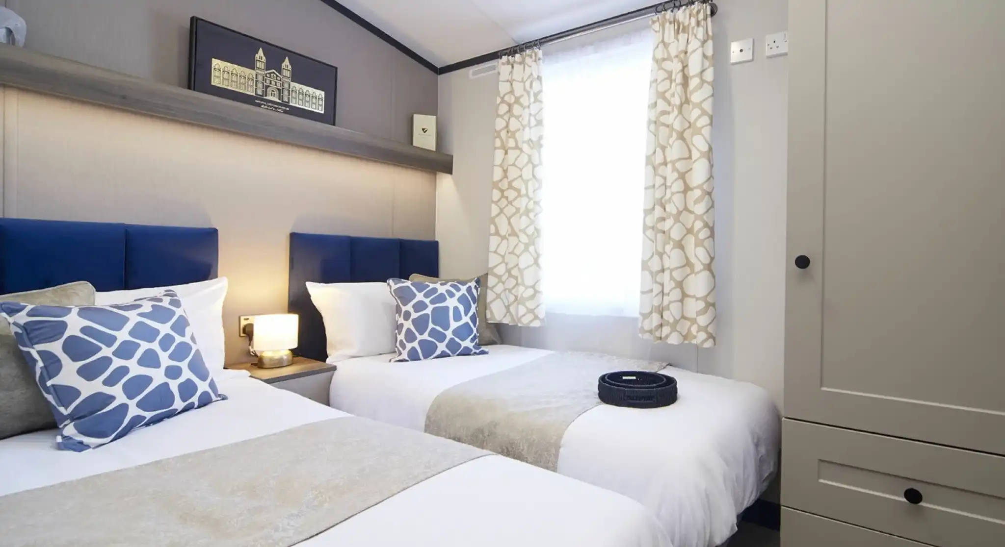 A stylish twin bedroom with twin beds with matching blue headboards, blue patterned cushions and white crisp linen. There is also a wardrobe and a window with patterned curtains allowing natural light.