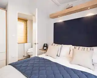 A bright master bedroom with double bed with blue accent cushions, bedspread and headboard, together with an en suite bathroom visible in the background with low-level WC.