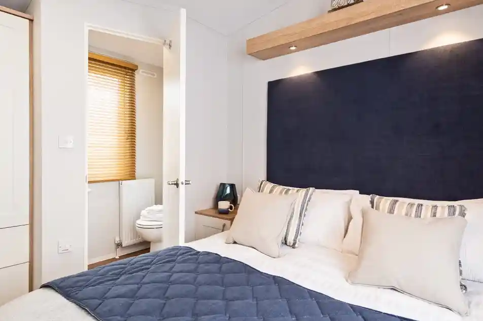 A bright master bedroom with double bed with blue accent cushions, bedspread and headboard, together with an en suite bathroom visible in the background with low-level WC.