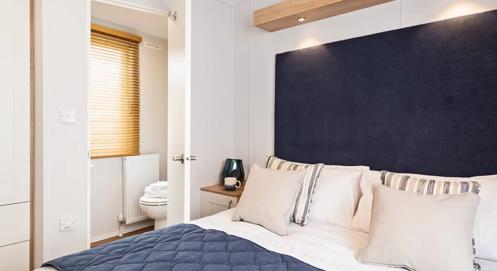 A bright master bedroom with double bed with blue accent cushions, bedspread and headboard, together with an en suite bathroom visible in the background with low-level WC.
