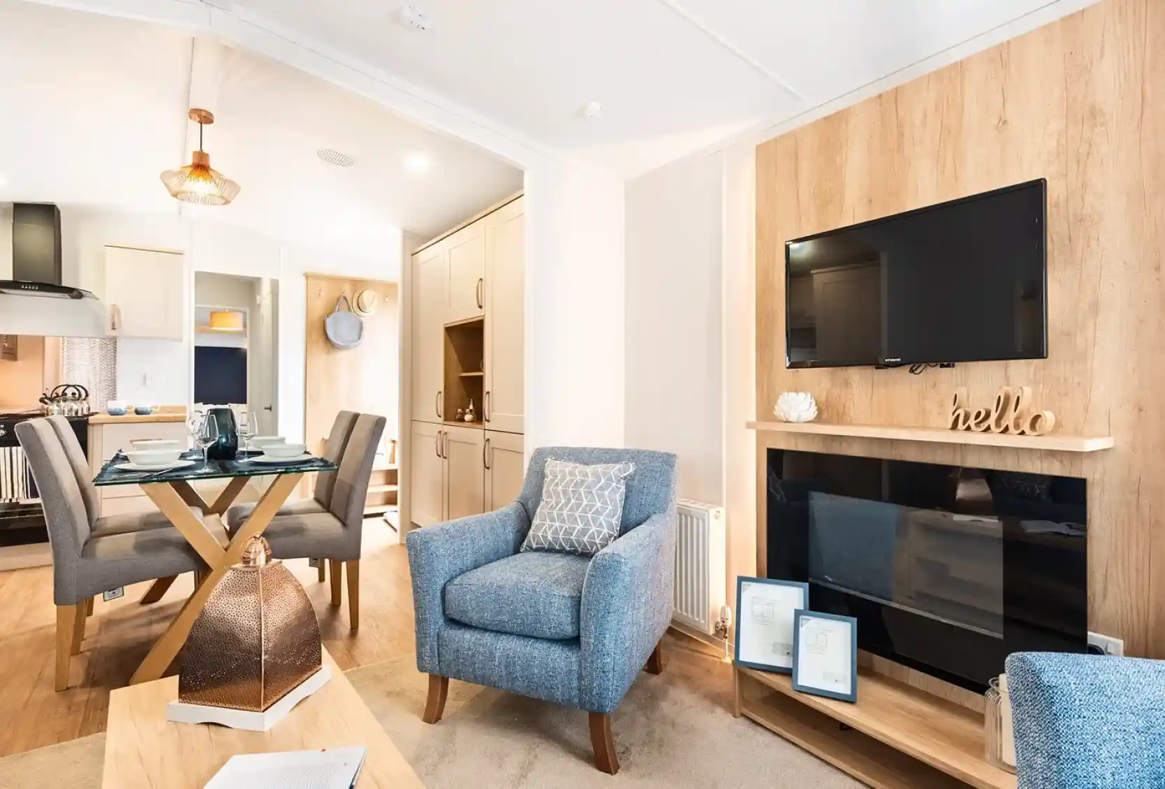 A feature wooden wall with electric fireplace and TV above, sat in a room with cosy armchairs and a dining room and kitchen in the background.