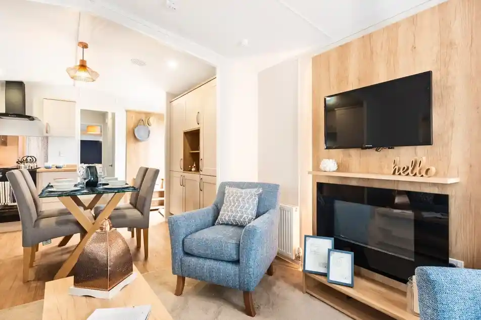 A feature wooden wall with electric fireplace and TV above, sat in a room with cosy armchairs and a dining room and kitchen in the background.