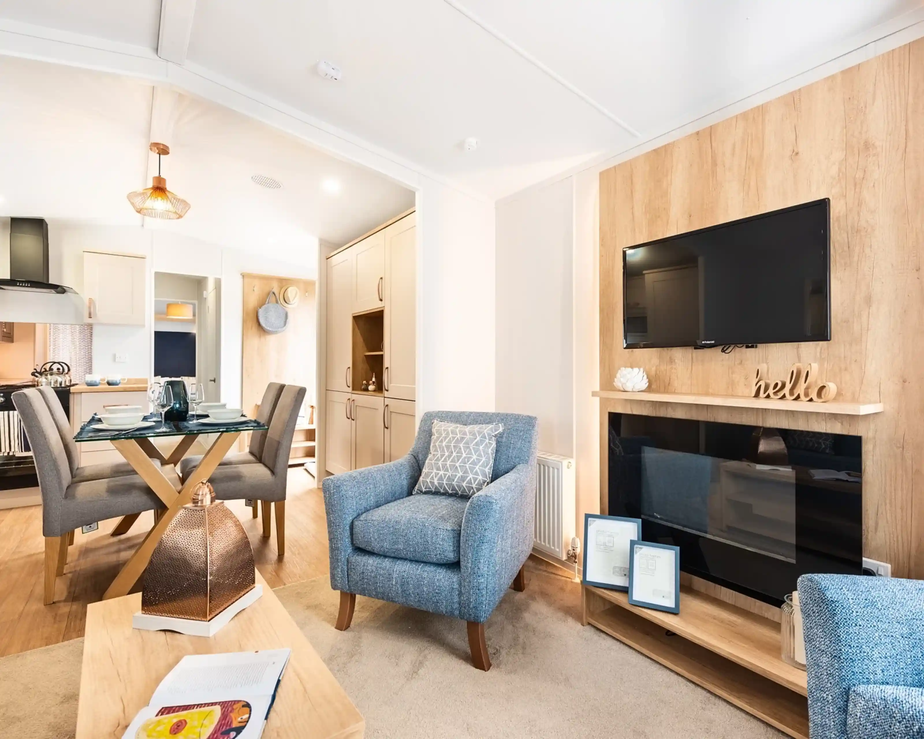 A feature wooden wall with electric fireplace and TV above, sat in a room with cosy armchairs and a dining room and kitchen in the background.